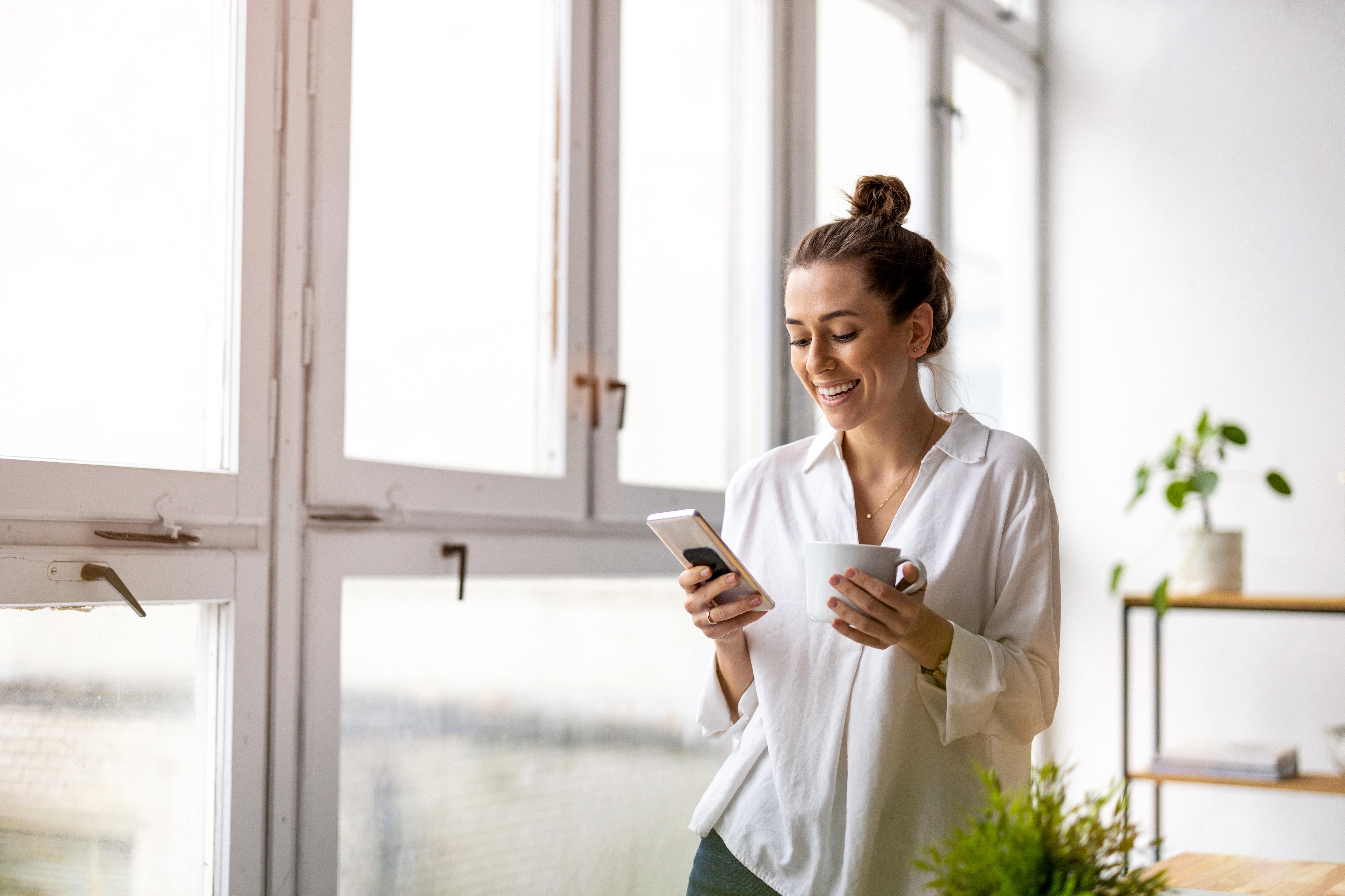 Lächelnde Frau hält ein Handy und eine Kaffeetasse.