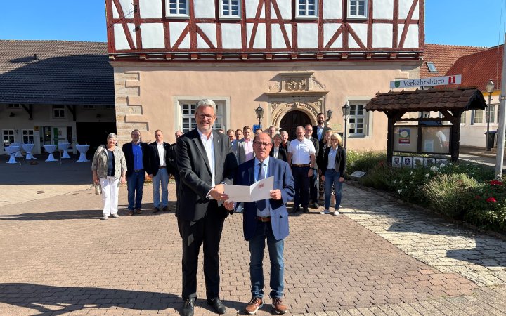 Zwei Personen stehend vor einer Gruppe von Personen bei Übergabe eines Dokuments.