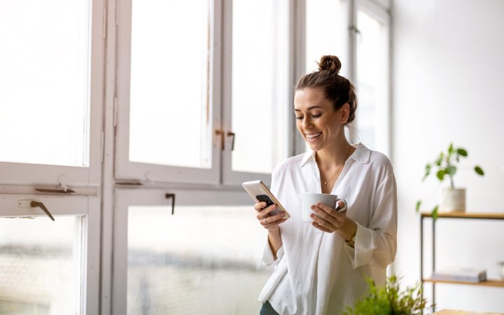 Lächelnde Frau hält ein Handy und eine Kaffeetasse.