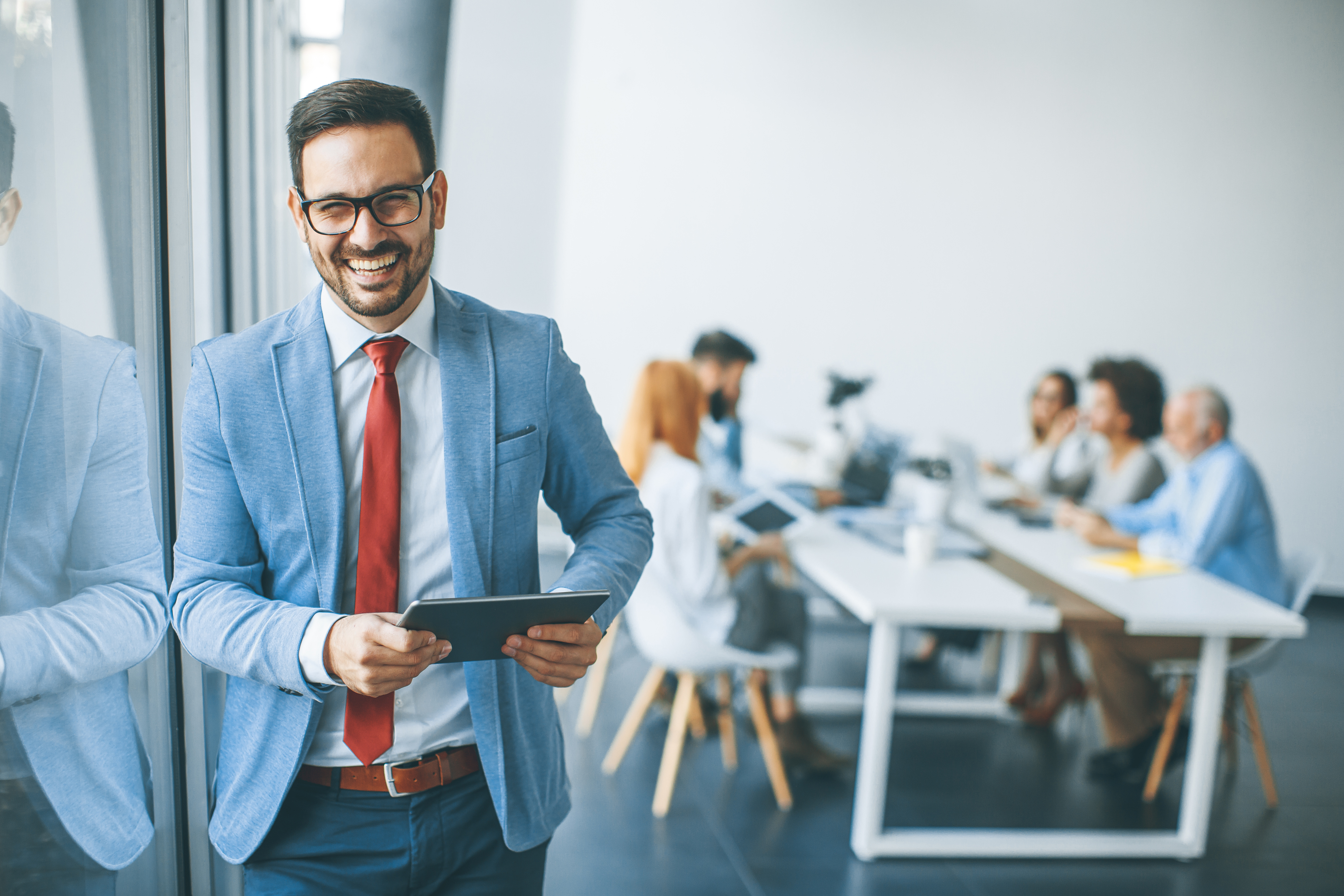 Lächelnder Mann hält ein Tablet in der Hand; im Hintergrund eine Gruppe von Personen.