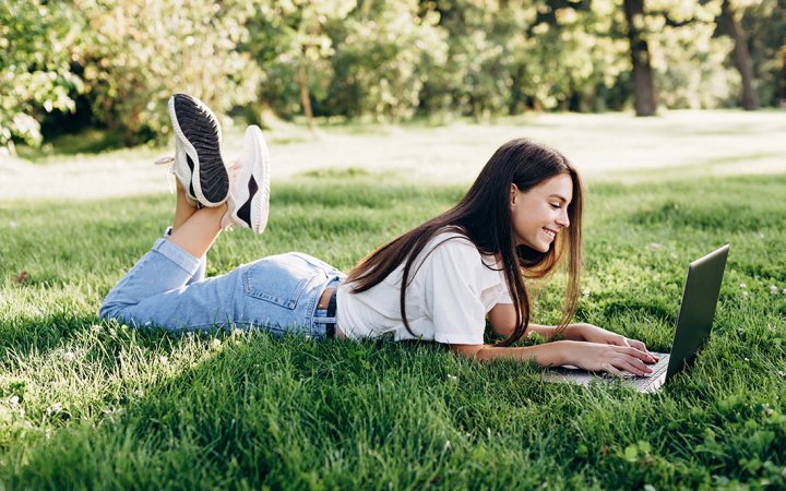 student girl with laptop outdoors. Smiling woman lying on the grass with a computer, surfing the Internet or preparing for exams. Technology, education and remote work concept. Soft selective focus.