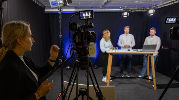 Eine Kamera filmt drei Personen bei einer Präsentation