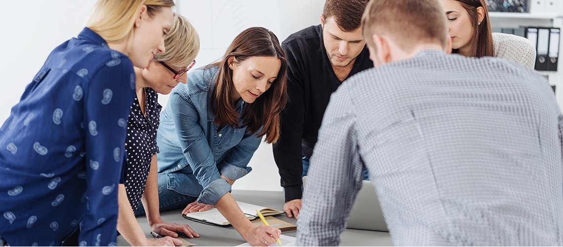 Sechs Personen gebeugt über Tisch; eine Frau zeigt auf Papiere auf dem Tisch